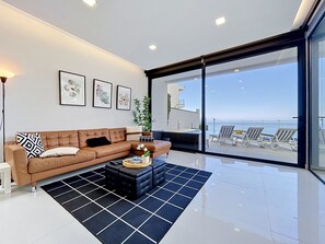 A modern living room with a brown leather sofa, a black ottoman, and a black and white rug. There are three framed artworks on the wall, a potted plant, and a view of the ocean through large glass doors. - Pearl of Madeira by Atlantic Holiday (Calheta)