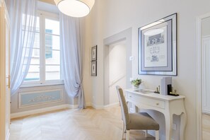 Ein Zimmer mit weißem Schreibtisch, einem Stuhl, einem gerahmten Bild und einem Fenster mit Vorhängen.