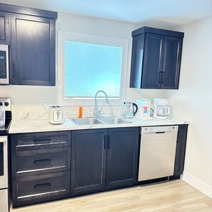 A modern kitchen with dark cabinets, a stainless steel dishwasher, a built-in oven, a microwave, a sink, and a window with blinds. - A cozy haven at the heart of the city and a minute drive to the airport (St. John's)