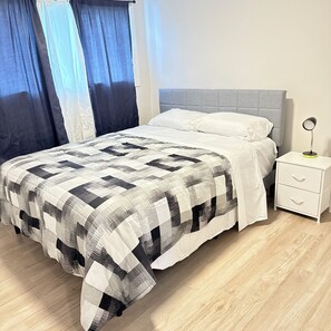 A bed with a checkered bedspread, a white pillow, a grey headboard, a bedside table with drawers, and a nightstand with a lamp.