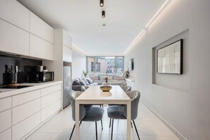 A modern kitchen with white cabinetry, a dining area with a white table and grey chairs, and a large window offering a city view. - Spacious 2BR in Psirri  2Baths  & Acropolis View (Athens)