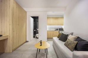 A modern living room with a wooden accent wall, a round coffee table, a sofa with cushions, and a kitchen in the background. - Generous Studio Syntagma | Worn Building Entrance (Athens)