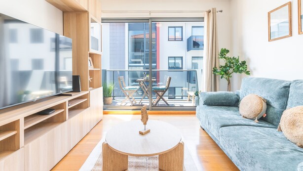A modern living room with a flat-screen TV, a round coffee table, a sofa, and a balcony with outdoor furniture.