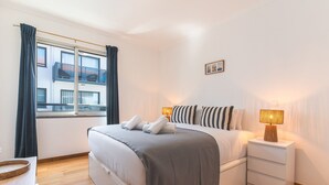 A neatly arranged bedroom with a bed, bedside tables, a window with curtains, and a view of buildings outside.