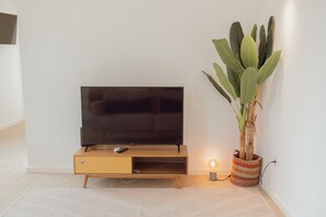 Ein modernes Wohnzimmer mit einem Flachbildfernseher auf einem Holzständer, einer kleinen Pflanze in einem gewobenen Korb und einer beleuchteten Stehlampe.