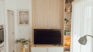 Sala de estar com televisão de tela plana, móvel para televisão em madeira, uma planta e uma estante com objetos decorativos.