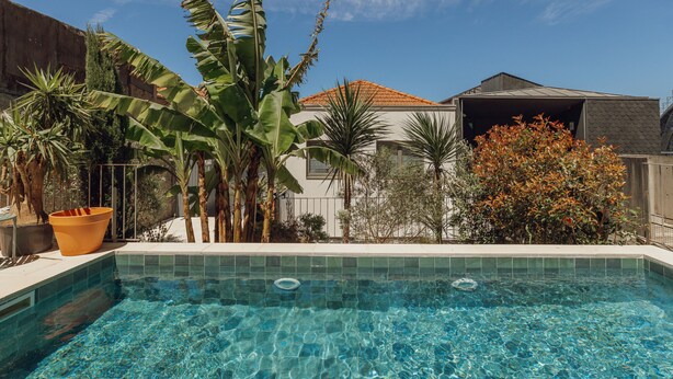 Uma piscina cristalina com borda azulejada, rodeada por plantas tropicais e um prédio residencial com telhado em cerâmica.