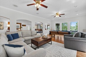 A spacious living room with a sofa, armchair, coffee table, and a pool table.