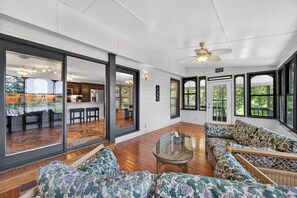 A spacious living area with large windows, a ceiling fan, and a view of the kitchen.