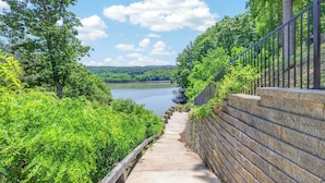 A concrete pathway leading to a body of water, surrounded by lush greenery and a stone wall with a metal railing. - Lakefront retreat with private dock, lake views, spacious decks, pool table, and sun room! (Pulaski)