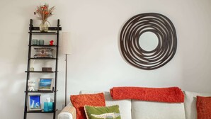 Un soggiorno con un divano bianco, cuscini decorativi rossi e verdi, una mensola in metallo nero con soprammobili e un’opera d’arte a parete circolare.