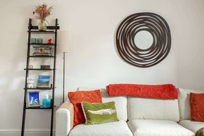 Ein Wohnzimmer mit einem weißen Sofa, roten und grünen Zierkissen, einem schwarzen Metallregal mit Dekorationsobjekten und einem runden Wandbild.