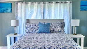 Un dormitorio con cama, cortinas, dos mesitas de noche con lámparas y ropa de cama estampada con motivos florales.