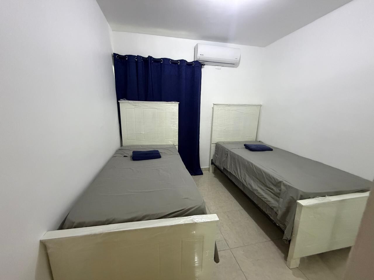 A small, single-room with two single beds, a window with curtains, an air conditioning unit, and a tiled floor.