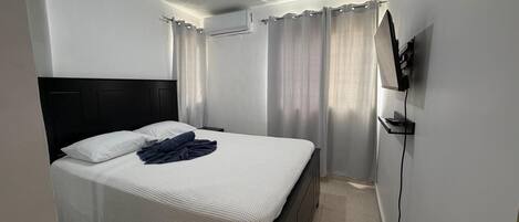 Un dormitorio con una cama, cortinas, un equipo de aire acondicionado y un televisor montado en la pared.