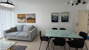 Uma sala de estar moderna com sofá, mesa de jantar de vidro e cadeiras pretas. Há duas obras de arte penduradas na parede e um ventilador de teto.