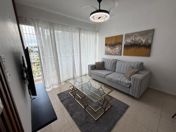 A living room with a sofa, a glass coffee table, a ceiling fan, and artwork on the wall.