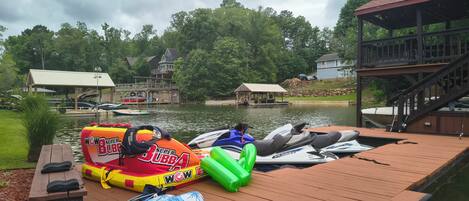 A wooden dock with inflatable boats and a kayak, a covered area, and a view of houses and trees across the water.