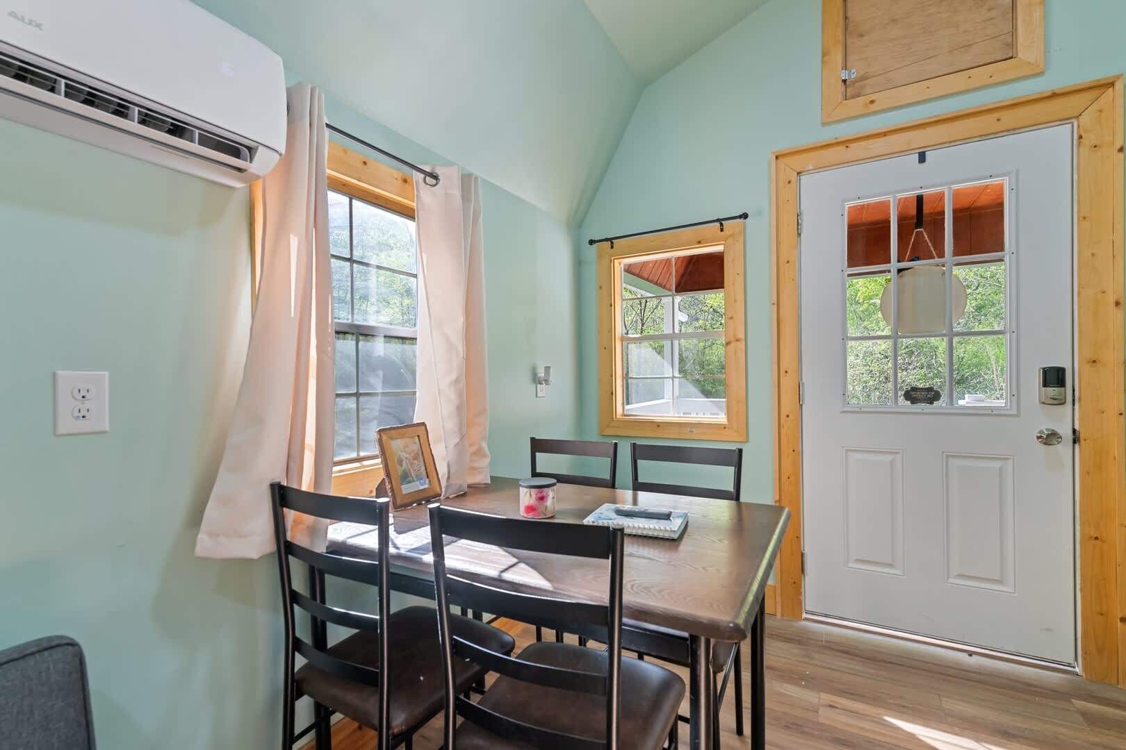 A room with a dining table, chairs, a window with curtains, a door, and an air conditioning unit.