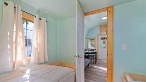 A room with two beds, a wooden door leading to a dining area, and a window with curtains. - Treehugger Tiny Home Cabin by River Near WCU (Cullowhee)