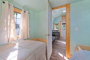 A room with two beds, a wooden door leading to a dining area, and a window with curtains. - Treehugger Tiny Home Cabin by River Near WCU (Cullowhee)