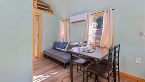 A room with a loft bed, a sofa, a dining table with chairs, and a window with curtains. - Treehugger Tiny Home Cabin by River Near WCU (Cullowhee)