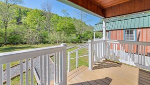 A wooden deck with a railing, a covered porch, and a view of a forested area. - Treehugger Tiny Home Cabin by River Near WCU (Cullowhee)