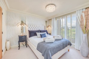 Ein Schlafzimmer mit einer gepolsterten Kopfteile, einem großen Bett mit vielen Kissen, Nachttischen, einer Kristallleuchter und Blick durch große Fenster.