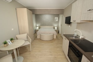 A compact bedroom with a bed, bedside tables, a small round table with chairs, a kitchenette with a sink, and a TV mounted on the wall. - Chloe by Frangki (Aegina)