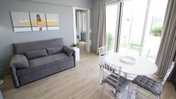 Una sala de estar con un sofá gris, una mesa redonda de comedor con sillas y un amplio ventanal con cortinas.
