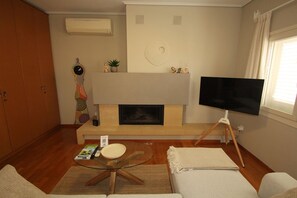 Ein Wohnzimmer mit Kamin, einem Fernseher auf einem Stativ, einer Couch, einem Couchtisch und einem Bücherregal.