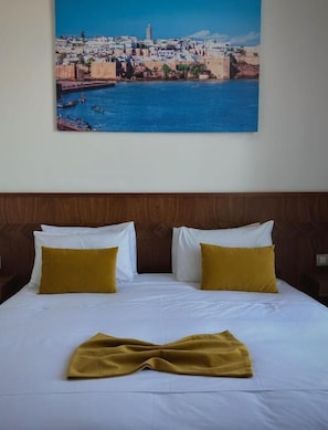 A hotel room with a bed, two pillows, a blanket, and a painting of a coastal cityscape. - Hôtel Majestic (rabat)