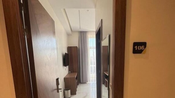 Pasillo de hotel con una puerta de madera, un cartel con un número y vistas a una habitación con cama y ventana.