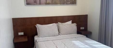 A bedroom with a bed, nightstands, a wall-mounted painting, and a window with curtains.