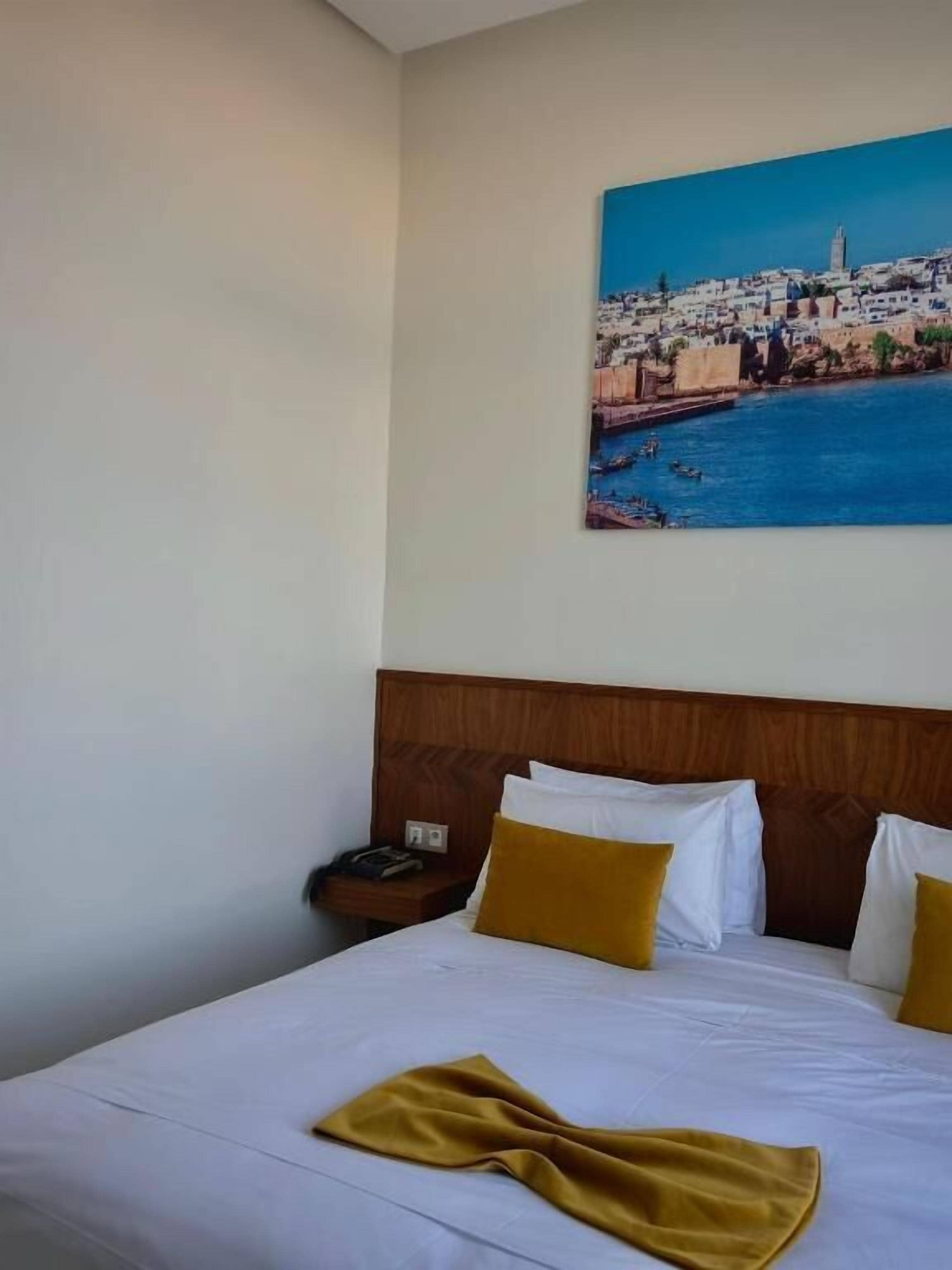 Hotelkamer met een bed, nachtkastje, schilderij van een kuststadsgezicht en een houten hoofdeinde.