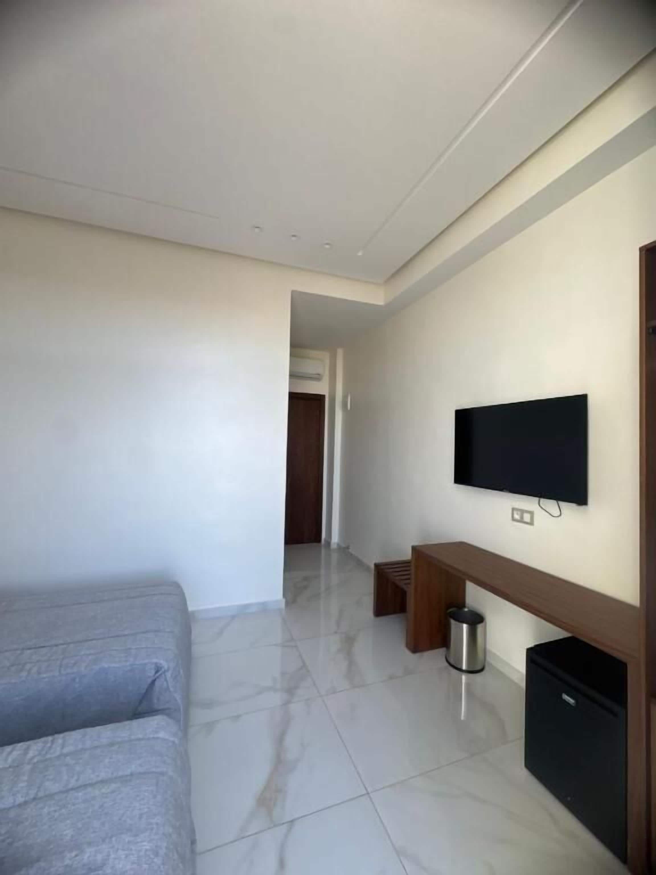 A hotel room with a bed, a TV mounted on the wall, a small wooden bench, a trash bin, and a storage unit.