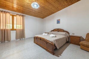 Ein Schlafzimmer mit Holzbalkendecke, einem Bett mit Holz Kopfteil, einem Nachttisch, einem Sessel und einem Fenster mit Vorhängen.