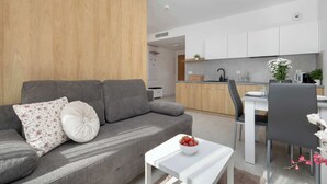 A modern living room with a grey sofa, white table, and a kitchen in the background. - Air Conditioned Apartment | Pool | Terrace | Parking (Mielno)