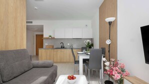 A modern living room with a grey sofa, a white coffee table with a bowl of fruit, a dining area with a white table and chairs, and a kitchen in the background. - Air Conditioned Apartment | Pool | Terrace | Parking (Mielno)