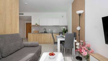 A modern living room with a grey sofa, a white coffee table with a bowl of fruit, a dining area with a white table and chairs, and a kitchen in the background.