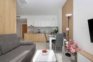 Ein modernes Wohnzimmer mit einem grauen Sofa, einem weißen Couchtisch mit einer Fruchtschale, einem Essbereich mit weißem Tisch und Stühlen sowie einer Küche im Hintergrund.