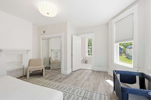 Una habitación con cama, una silla, un ventanal con persianas, una puerta que da a otra habitación y una lámpara en el techo.