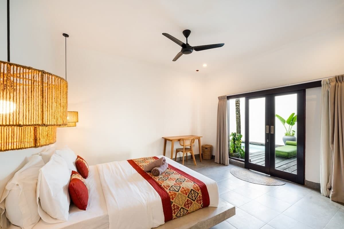 A modern hotel room with a bed, a ceiling fan, a table, and a sliding glass door leading to an outdoor area.