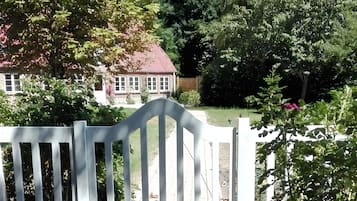 Ein weißer Holzzaun, ein Ziegelhaus mit rotem Dach und üppiges Grün im Hintergrund.