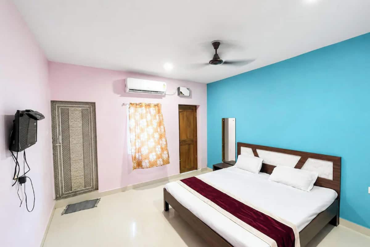 A bedroom with a bed, a ceiling fan, an air conditioning unit, a television, and a door.