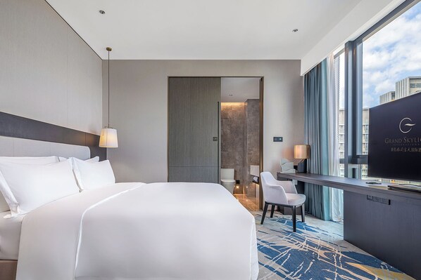 A hotel room with a large bed, a desk with a television, and a view of the city.