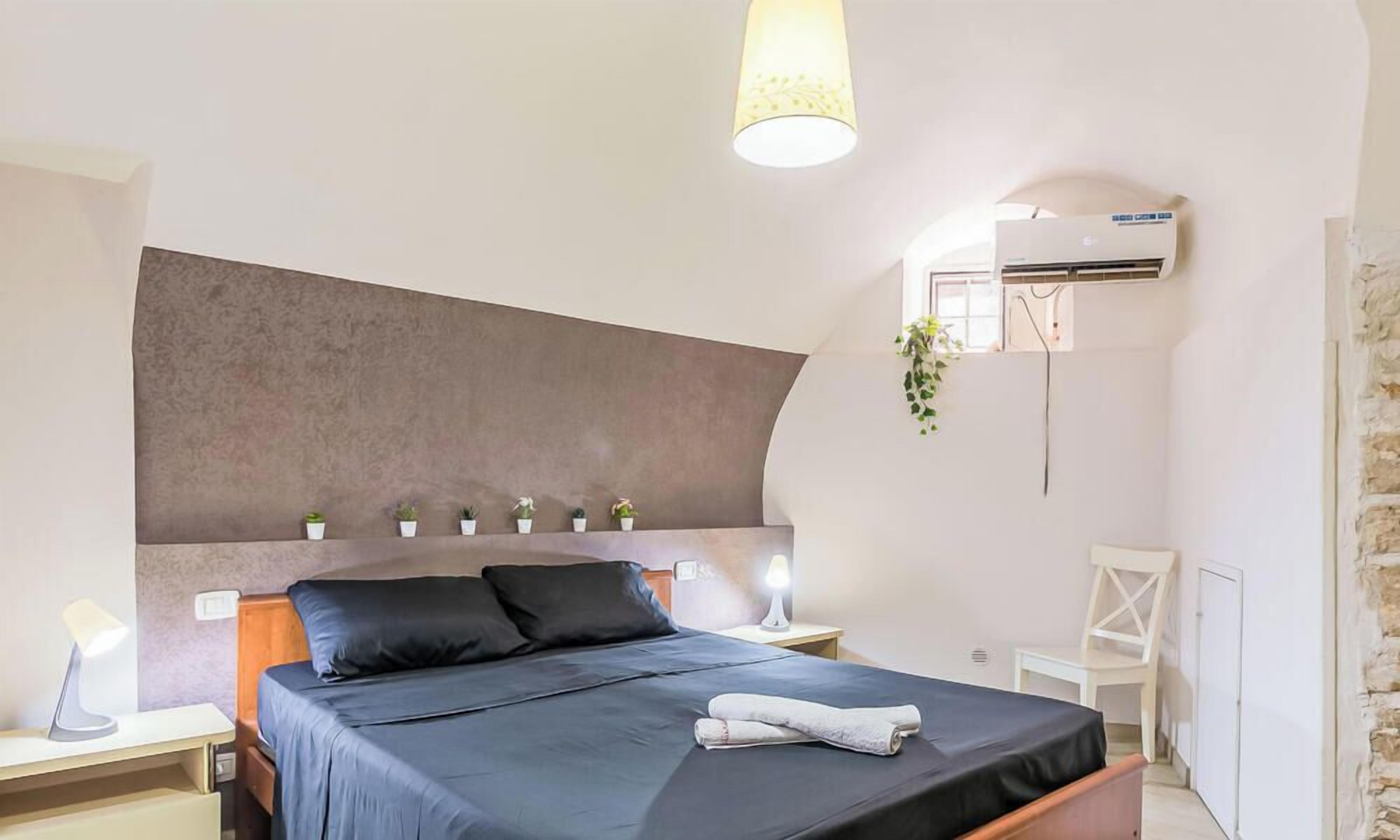 A bedroom with a bed, bedside lamps, a wall-mounted air conditioner, a small plant, and a white chair.