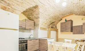 Une cuisine avec des murs en pierre, des meubles en bois, un poêle et un coin repas avec des chaises blanches.