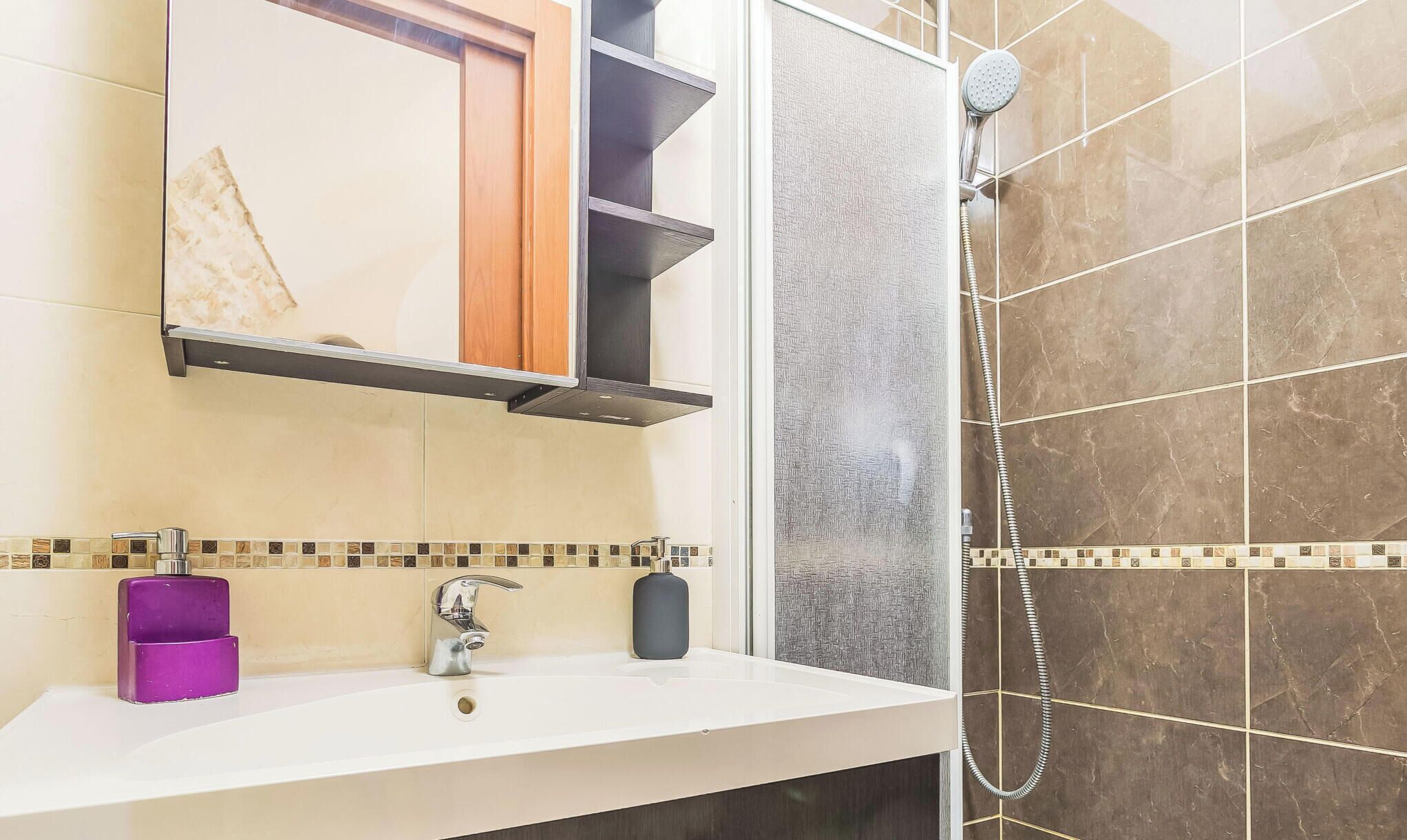 Une salle de bain avec un lavabo blanc, un distributeur de savon violet, un miroir avec un cadre en bois et une douche avec une porte en verre.