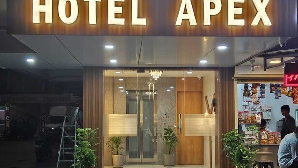 Ein Hotel Eingang mit einem Schild mit der Aufschrift 'ROOMS HOTEL APEX', Pflanzen in Töpfen und einer draußen sitzenden Person.